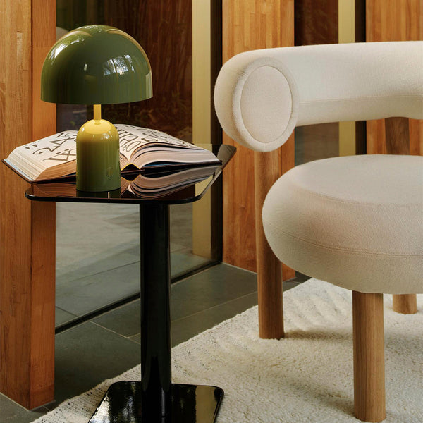 Green dome lamp on a sleek black table beside a soft white chair, accented by warm wooden elements and a textured rug.