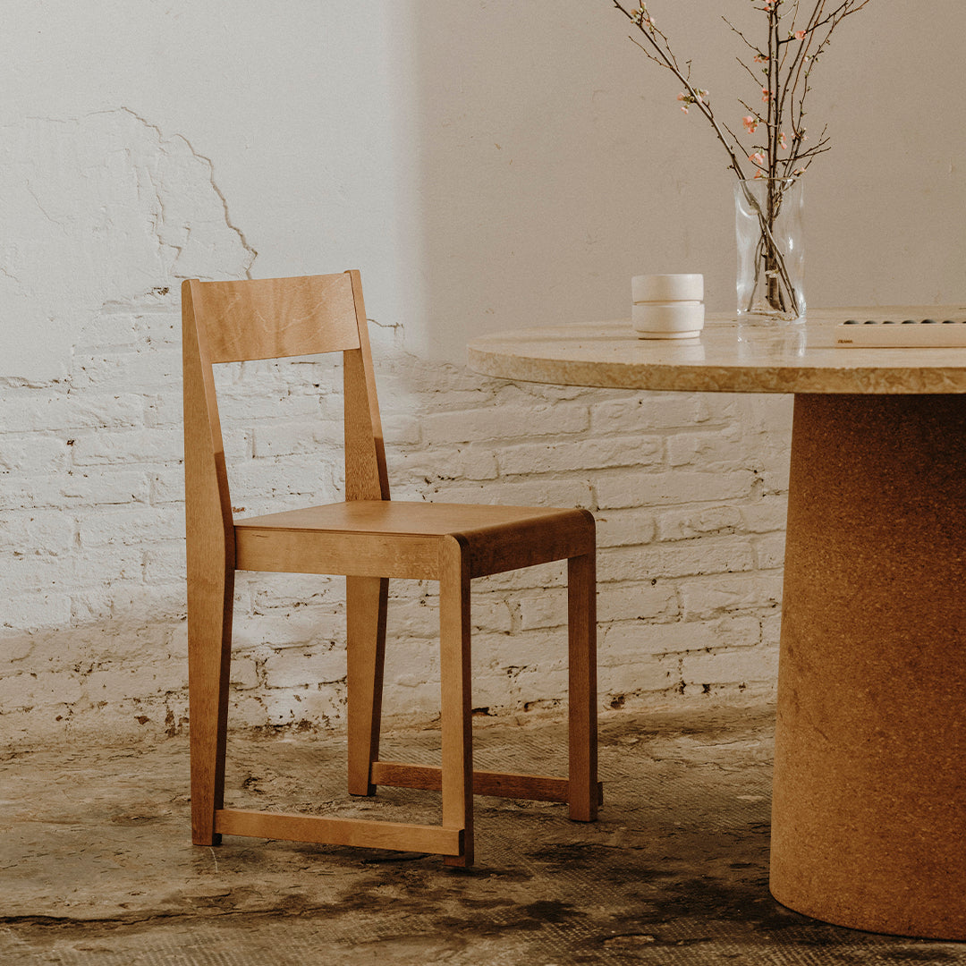 Light wooden chair next to a beige round table, set against a textured white brick wall. Minimalist design.