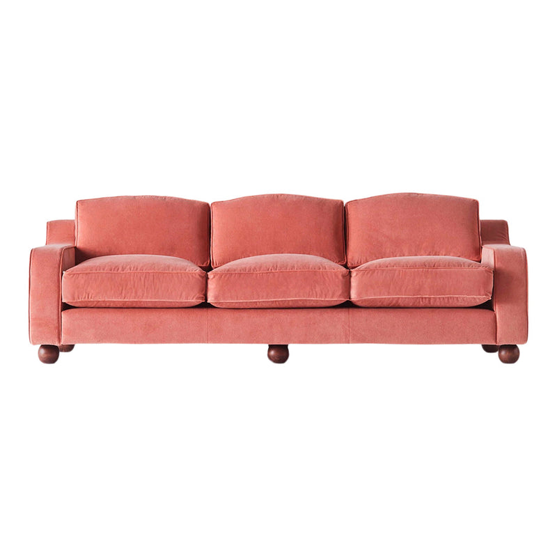 Soft coral velvet sofa with rounded cushions and dark wooden feet. Modern design ideal for cozy living spaces.