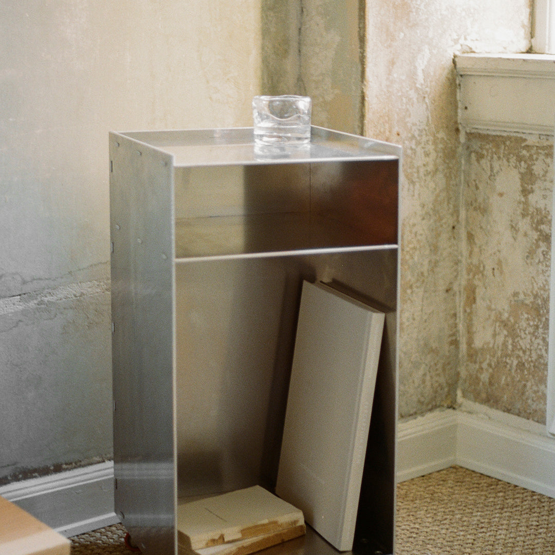 Sleek stainless steel side table with shiny surface, showcasing minimalist design against a textured wall backdrop.