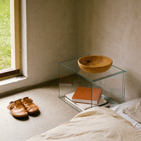 Transparent glass side table with a natural wood bowl, earthy tones, and light neutrals in a serene bedroom setting.