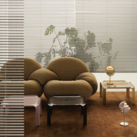 Brown tufted sofa with subtle pattern, pastel pink and clear tables, natural greenery, and soft white blinds in modern setting.