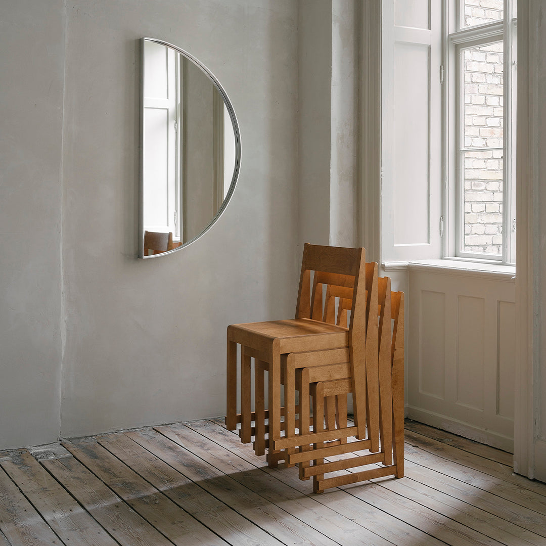 Modern wooden chairs in natural wood finish, stacked neatly against a light gray wall with a circular mirror.