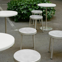 White circular tables and stools with sleek silver legs, set against a green backdrop. Modern and minimalist design.
