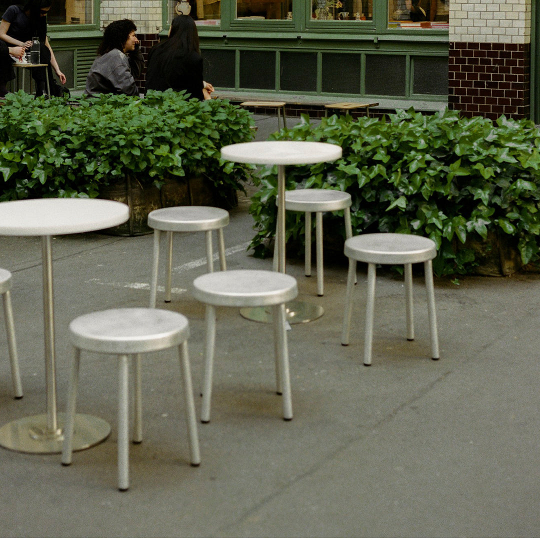 Modern white tables and stools with sleek metal legs, set against lush green foliage and an urban backdrop.