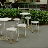 Modern white tables and stools with sleek metal legs, set against lush green foliage and an urban backdrop.