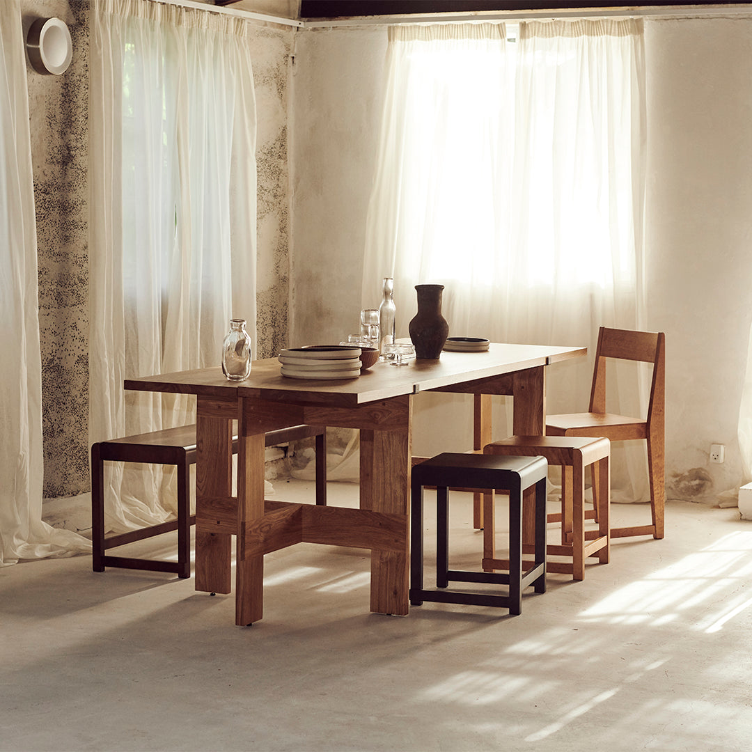 Natural wood table with light brown chairs, black stools, and neutral-toned curtains in a softly lit interior.