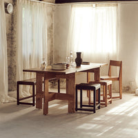Natural wood table with light brown chairs, black stools, and neutral-toned curtains in a softly lit interior.