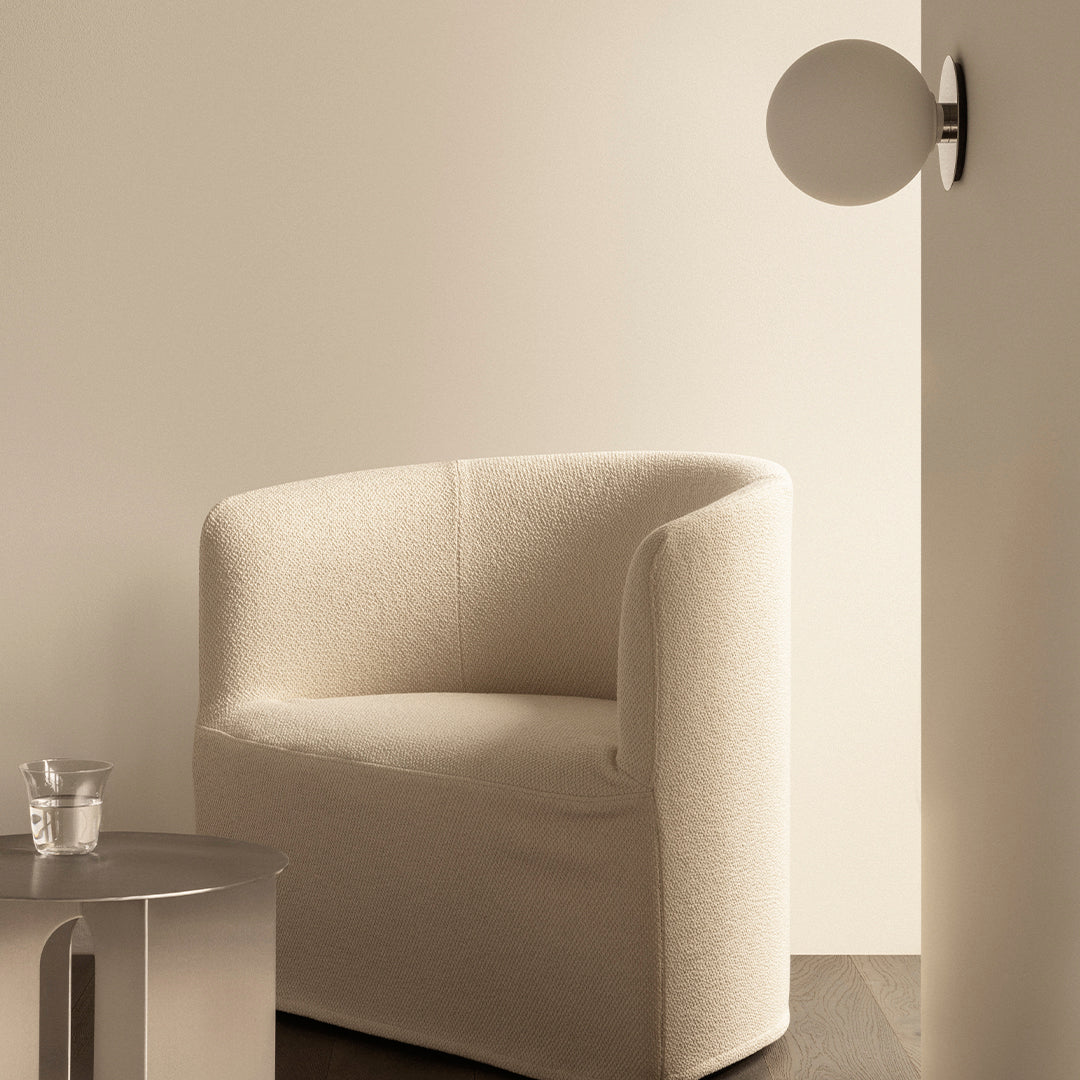 Beige textured armchair against a soft, light wall, complemented by a sleek silver side table and glass.