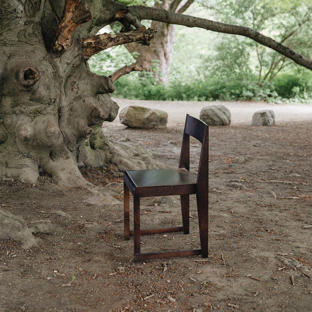 "Minimalist dark wood chair with clean lines, set against a natural outdoor backdrop of earthy tones."