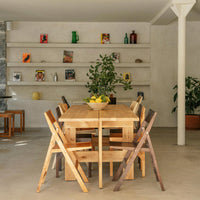 Wooden dining table with light and dark brown folding chairs, complemented by greenery and neutral-colored walls.
