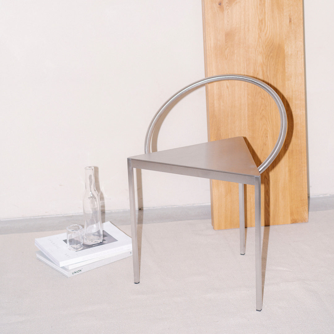 Modern silver triangle chair with circular backrest, paired with a natural wood backdrop. Minimalist design.
