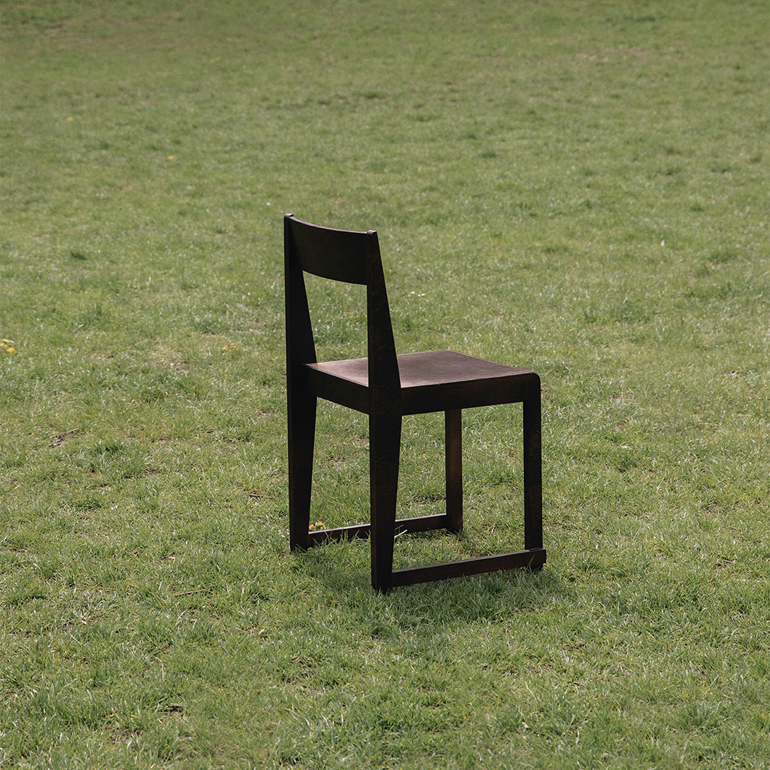 Black wooden chair with a simple design, featuring a smooth seat and straight lines, set against a grassy background.