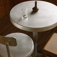 Round silver table with smooth finish, paired with a light wooden chair, creating a minimalist and modern aesthetic.