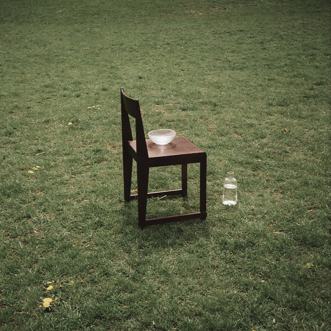 Dark brown wooden chair on green grass with a clear bowl and a transparent bottle nearby. Simple, minimalist design.