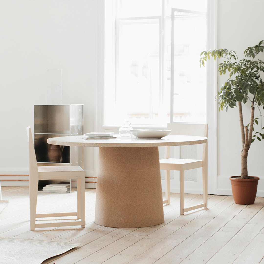 Light wood dining table with a round cork base, paired with white chairs in a bright, airy room with natural light.