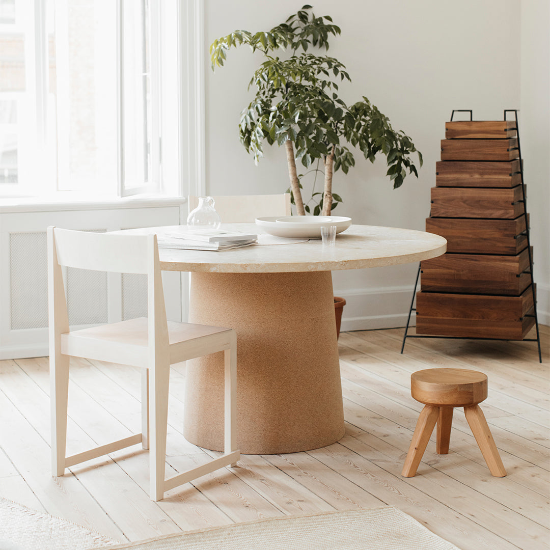 Modern round white table with a textured beige base, paired with light wood chairs and a wooden stool in a neutral interior.