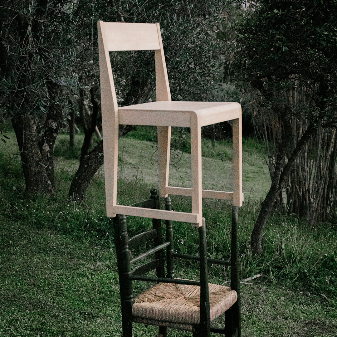 Light-colored wooden chair positioned atop a black chair, set against a natural green landscape.