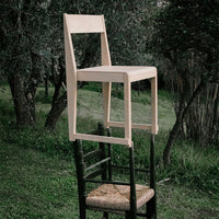 Light-colored wooden chair positioned atop a black chair, set against a natural green landscape.