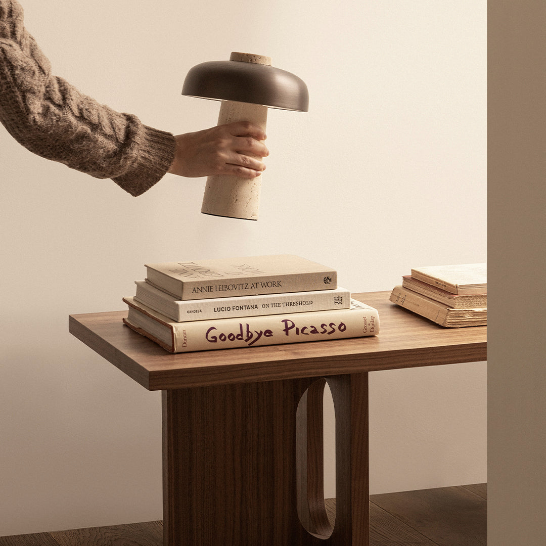 Hand holding a black and beige lamp above stacked books on a wooden table, showcasing warm neutrals and natural textures.