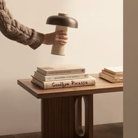 Hand holding a black and beige lamp above stacked books on a wooden table, showcasing warm neutrals and natural textures.