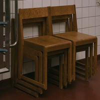 Stacked wooden chairs with a warm brown finish, featuring a smooth texture and minimalist design against a tile backdrop.