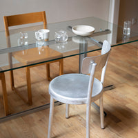 Gray velvet chair beside a glass table, highlighted by wooden chair and minimalist dishware on a light wood floor.
