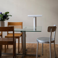 Modern dining table with glass top, wooden and silver chairs, and white lamp; neutral wall and warm wooden floor.