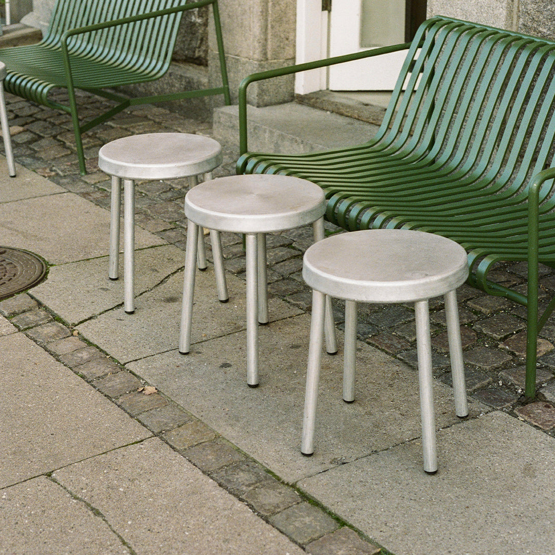 Three round, gray stools with silver legs beside a green, striped bench on a stone pavement.