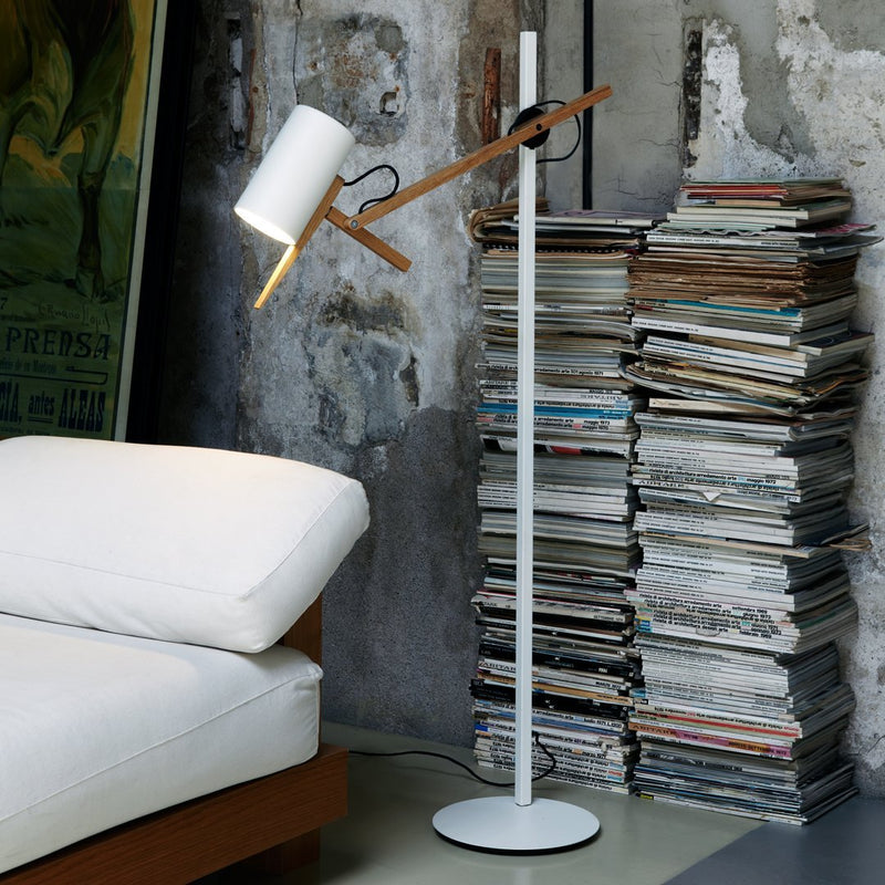 White floor lamp with wood accents against a textured gray wall, complemented by a stack of magazines.