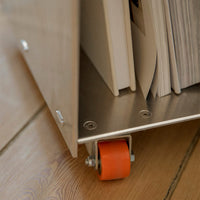 Close-up of a modern storage cart with a sleek metallic finish and bright orange wheels, set against a wooden floor.