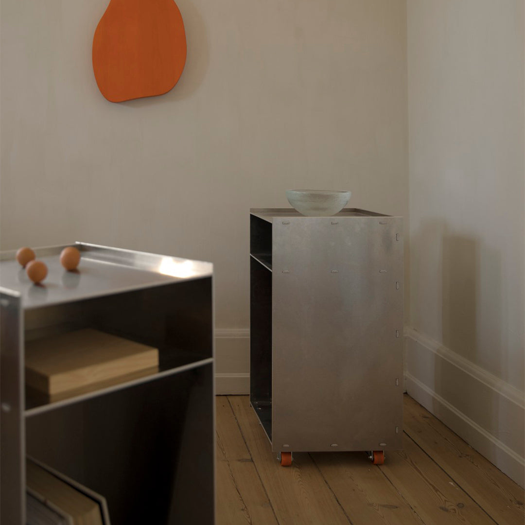 Metallic silver storage cart on wheels with orange accents in a minimalist, neutral-toned room.