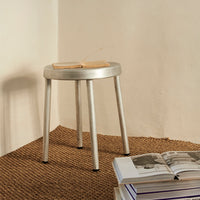 Silver minimalist stool with a round top and sleek legs, placed on a textured brown rug. Neutral backdrop.