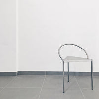 Sleek silver metal chair with a minimalist triangular design, set against a clean white wall and gray tiled floor.
