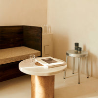 "Marble table with circular top and cork base, complemented by a sleek silver stool in a warm-toned interior."