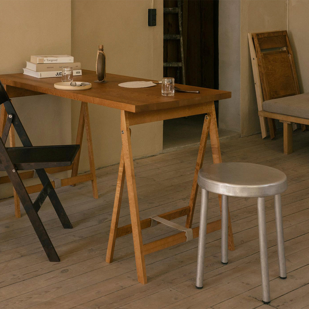 Wooden folding table in warm brown tones, paired with a sleek silver metal stool on light wooden flooring.