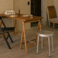 Wooden folding table in warm brown tones, paired with a sleek silver metal stool on light wooden flooring.