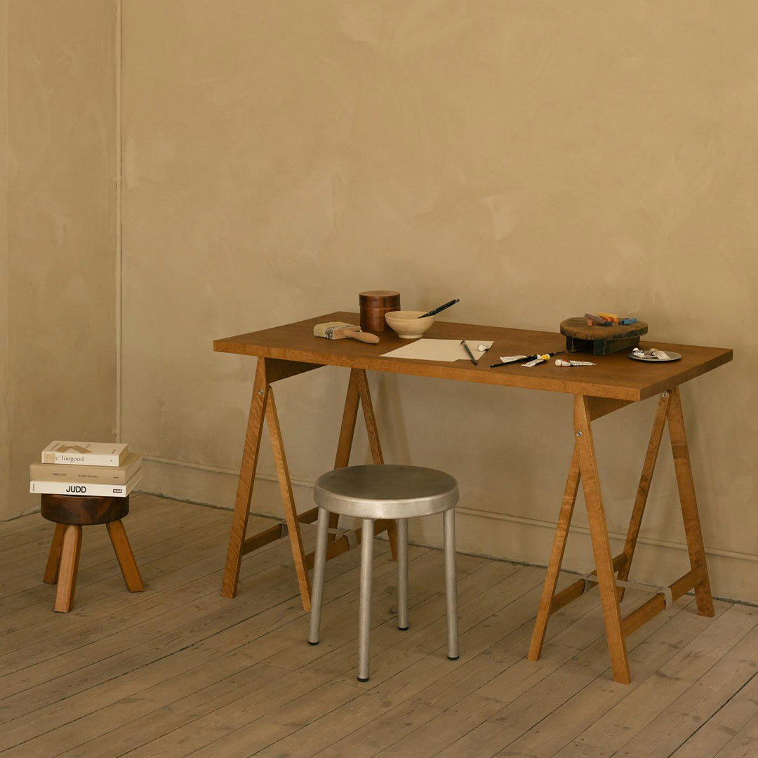 Wooden desk in warm brown tones with a sleek silver stool against a textured beige wall. Minimalist design elements.