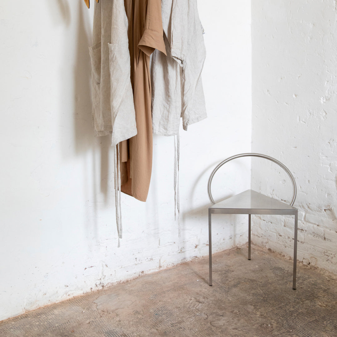 Minimalist silver chair against a textured beige floor, with neutral-toned hanging garments.