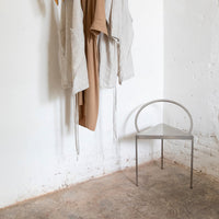Minimalist silver chair against a textured beige floor, with neutral-toned hanging garments.