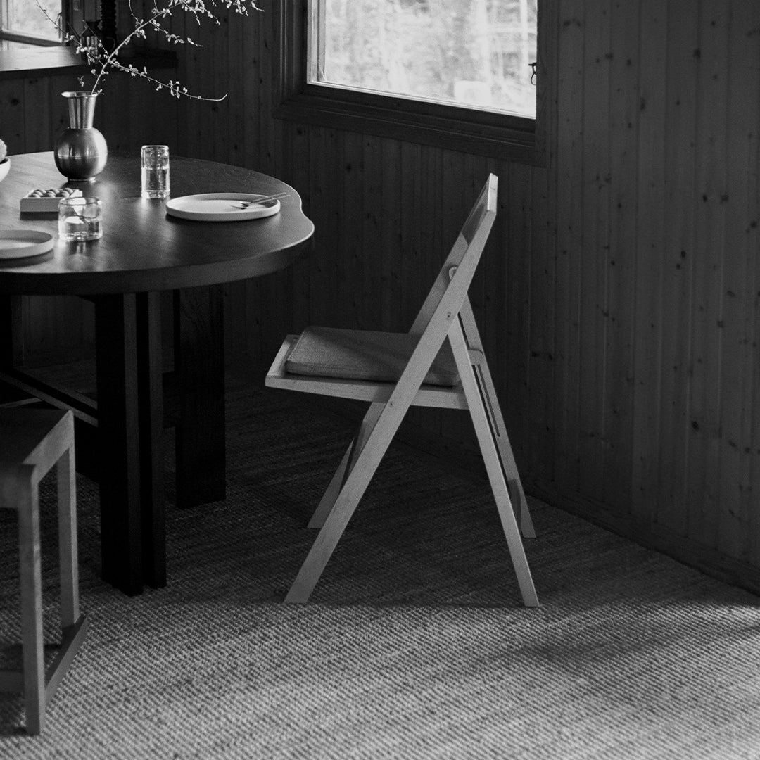Black and white image of a modern foldable chair with simple lines, placed by a round dark wooden table and textured rug.