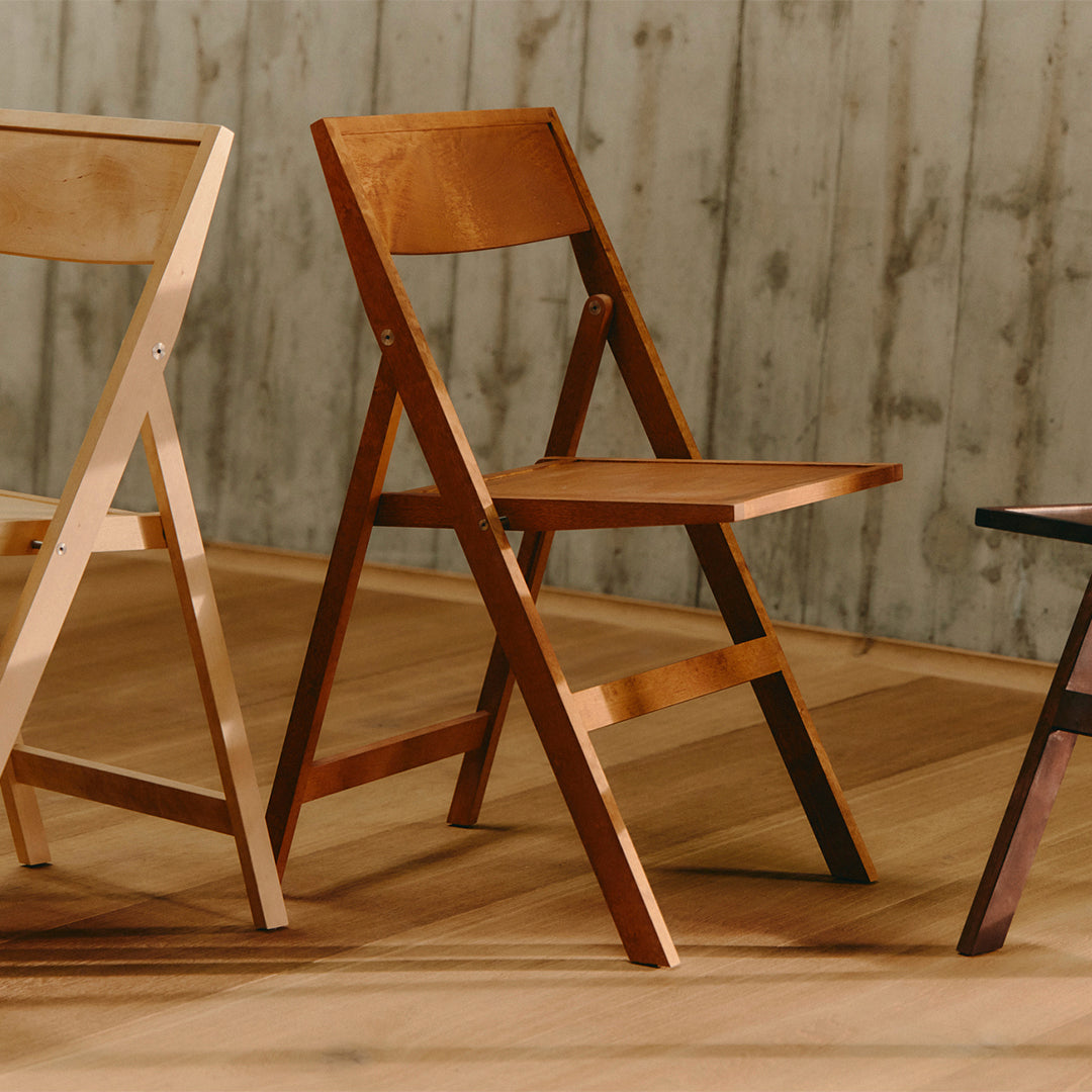 Three wooden folding chairs in light beige, warm medium brown, and dark espresso, set against a textured wooden backdrop.