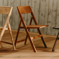 Three wooden folding chairs in light beige, warm medium brown, and dark espresso, set against a textured wooden backdrop.