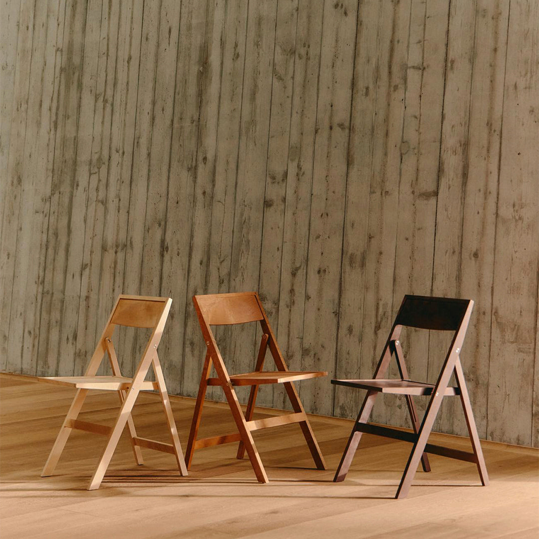 Three wooden folding chairs in natural, light orange, and black finishes, set against a textured gray wall.