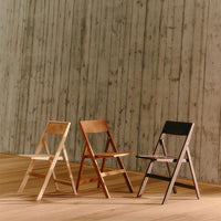 Three wooden folding chairs in natural, light orange, and black finishes, set against a textured gray wall.