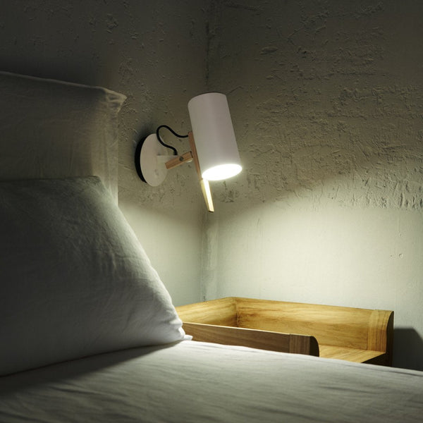 White cylindrical lamp with wooden accent on a textured gray wall, illuminating a light-colored bed and wooden nightstand.