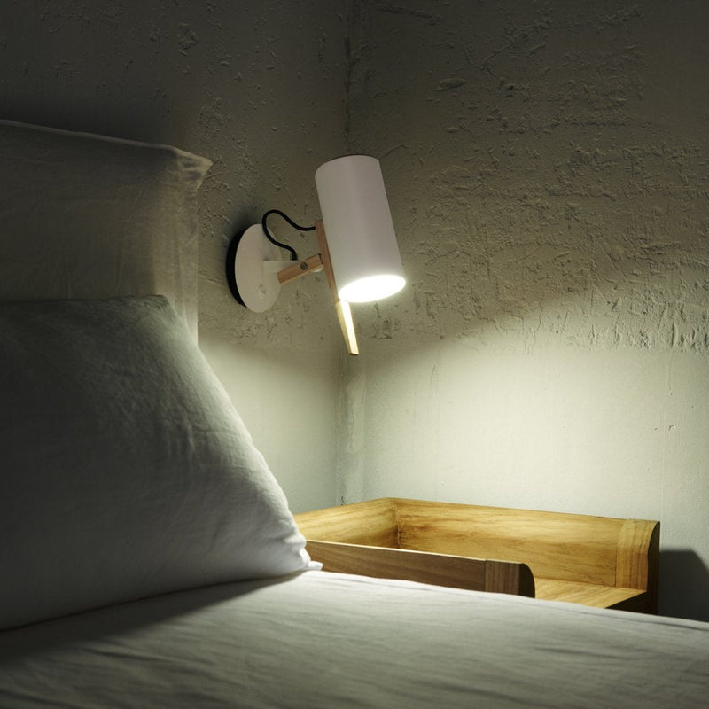 White cylindrical lamp with wooden accent on a textured gray wall, illuminating a light-colored bed and wooden nightstand.