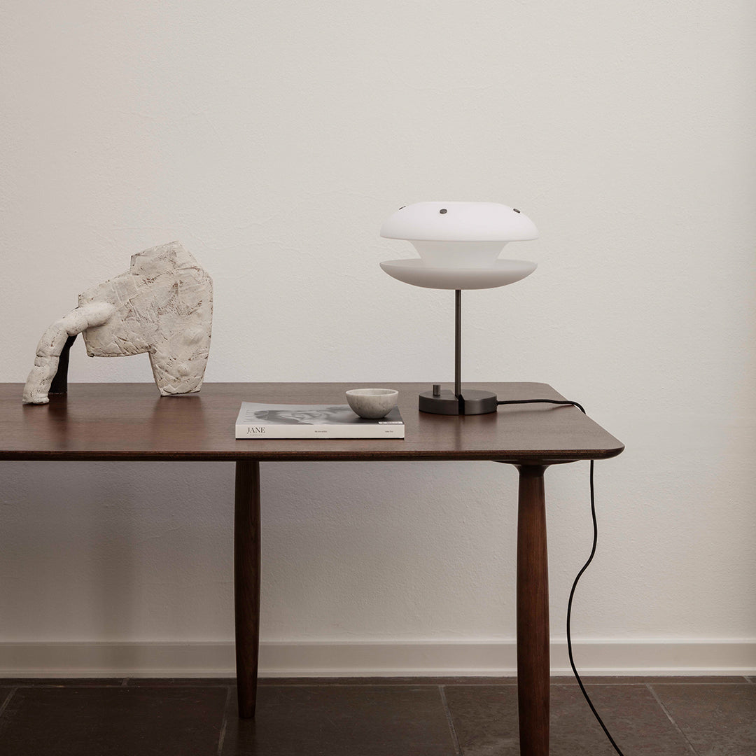 White modern lamp on a walnut table, accented by a light gray sculpture and dark tile flooring. Minimalistic style.