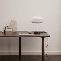 White modern lamp on a walnut table, accented by a light gray sculpture and dark tile flooring. Minimalistic style.