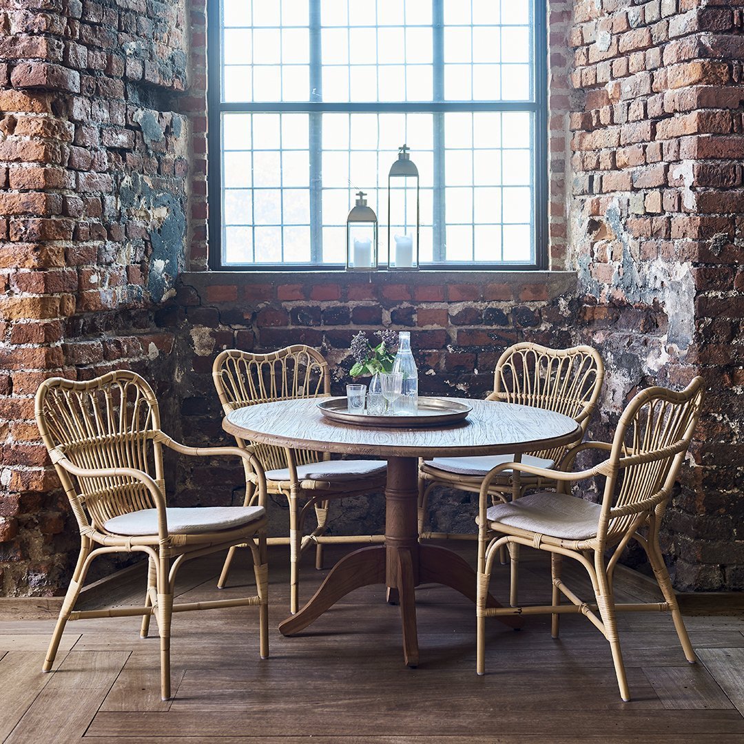 Rattan dining set with natural tones, featuring a round white tabletop and cream cushions against a rustic brick backdrop.